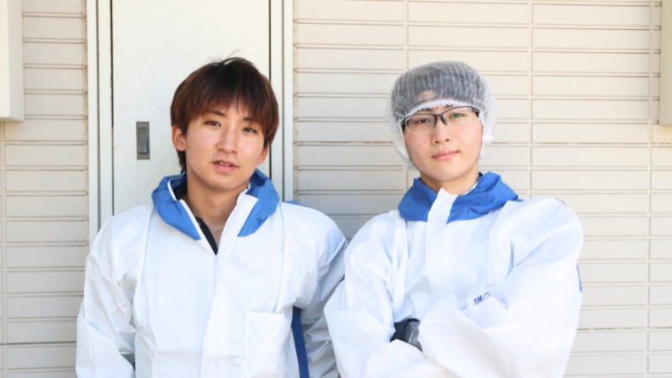 白い防護服を着用した2名の若い男性スタッフが建物前に並んで立っている様子