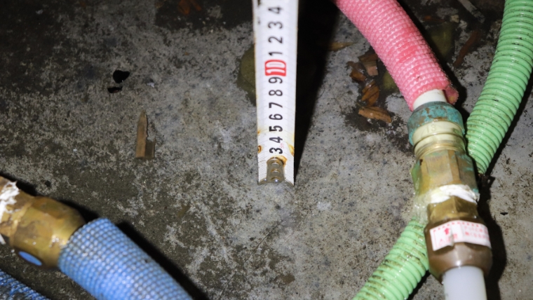 自社スタッフが撮影した床下浸水現場。コンクリート基礎面にカビが発生しており、給水・排水配管とともにメジャーで浸水深さを実測している様子