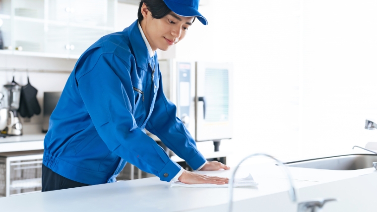 青い作業服と帽子を着用した清掃員がキッチンのカウンターを拭き掃除している様子