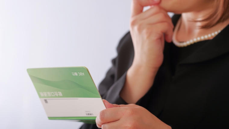 黒い喪服とパールネックレスを着用した女性と思われる人物が、銀行の通帳と思われる冊子を手に持ち考え込んでいるイメージ。孤独死後の処理費用負担を誰が担うか思案する状況を想起させる