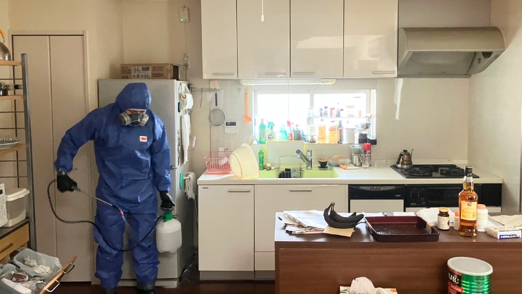 青い防護服と3M製防毒マスクを着用した自社スタッフが、生活用品が残るキッチンで噴霧器を使って実際に消毒・除菌作業を行っている様子
