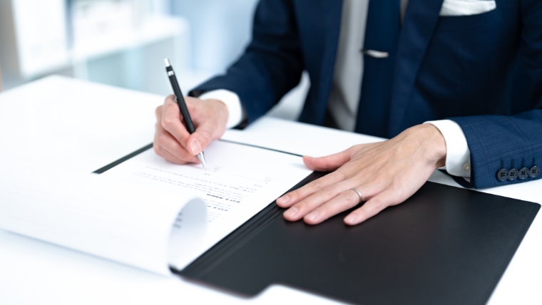 スーツ姿と思われる人物が書類にペンで記入しているイメージ。孤独死後に必要な各種手続きや書類作成を想起させる