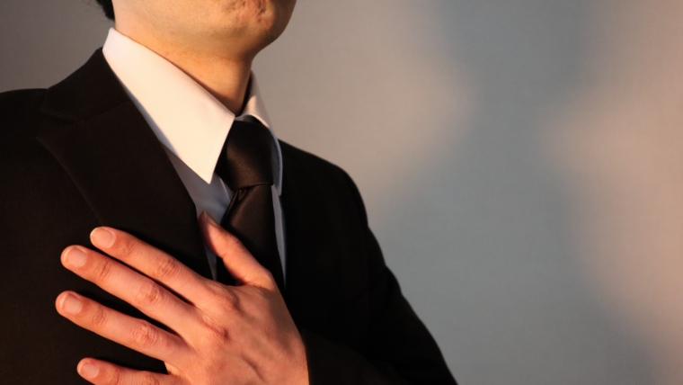 黒いスーツと黒ネクタイ姿と思われる人物が胸に手を当てているイメージ。孤独死後の処理における遺族や関係者の責任と役割を想起させる