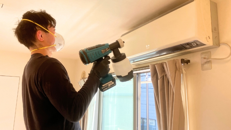 マスクと黒いゴム手袋を着用した自社スタッフが、電動スプレーガンを使ってエアコン周辺に防カビ処理を実際に施工している様子