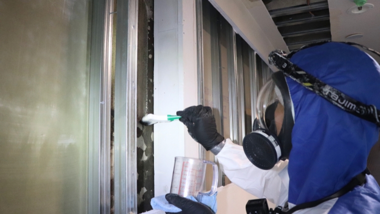 青い防護服と防毒マスクを着用した自社スタッフが、計量カップに入れた薬剤をハケで窓サッシのカビに実際に塗布している様子