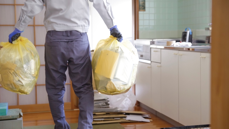 作業服と思われる服装の人物が青いゴム手袋を着用し、室内で黄色いゴミ袋を両手に持って運んでいるイメージ。孤独死後の遺品整理・相続手続きに伴う片付け作業を想起させる