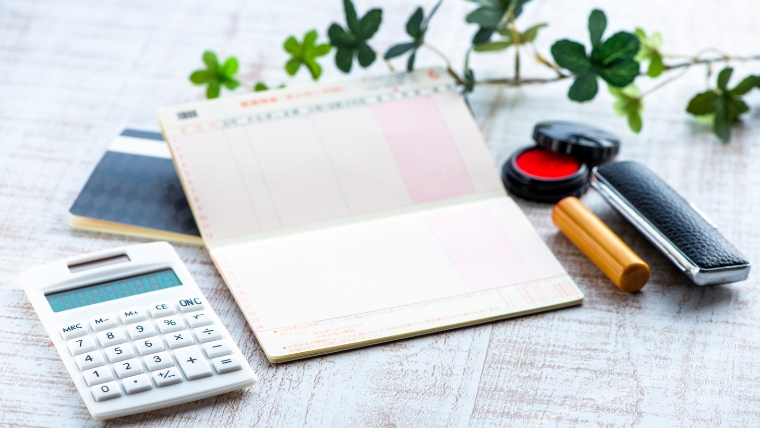 通帳と思われる冊子・電卓・印鑑・朱肉が机に並んでいるイメージ。孤独死後の生活関連手続きに関する各種書類整理を想起させる