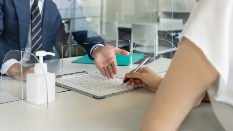 アクリル板越しにスーツ姿と思われる人物と依頼者が契約書に署名しているイメージ。身寄りのない孤独死後に行政や専門家へ手続きを委任する状況を想起させる