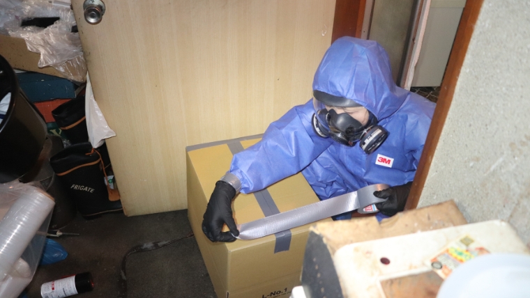 青い防護服と3M製防毒マスクを着用した作業員と思われる人物が、室内で段ボール箱をガムテープで封をしているイメージ。賃貸物件での孤独死後に行われる特殊清掃・原状回復作業を想起させる