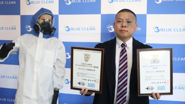 ブルークリーンののぼりを背景に、スーツ姿の男性が額入り認定証2枚を手に持ち、隣に防護服とガスマスク姿の人物が並んでいる様子