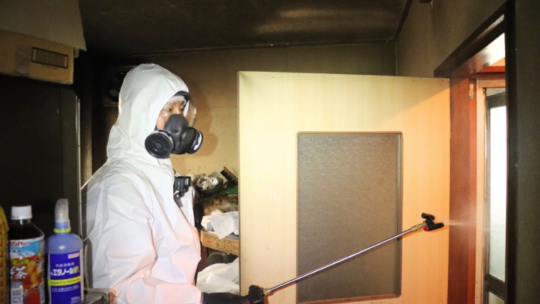 白い防護服とガスマスクを着用した特殊清掃員が室内でスプレーガンを使い薬剤を散布している様子