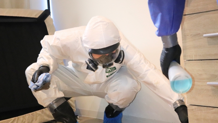 白い防護服と防毒マスクを着用した特殊清掃員が青い液体の入った容器を持ちながら床付近で作業している様子