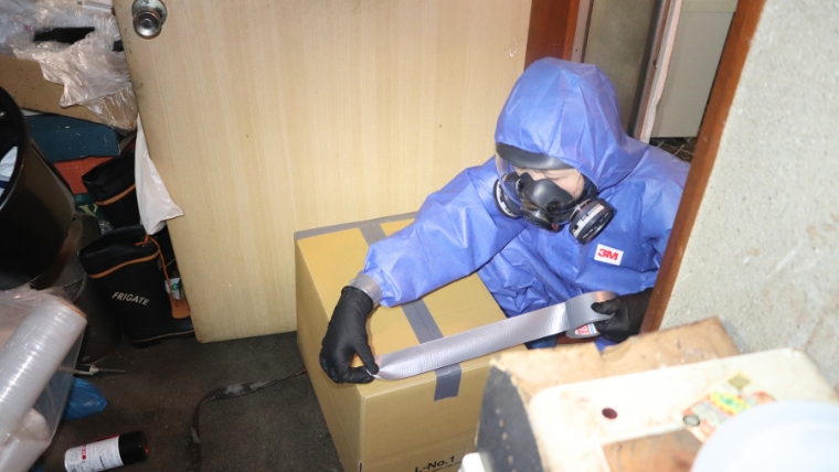 青い防護服と3M製ガスマスクを着用した特殊清掃員1名がダンボール箱をテープで梱包する様子