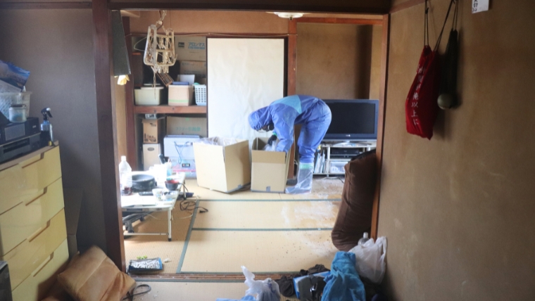 青い防護服を着用した特殊清掃員が和室でダンボール箱を扱いながら作業している様子