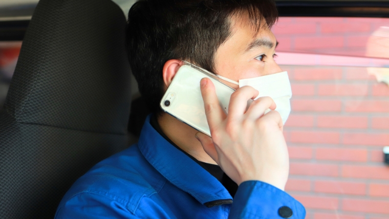 青いユニフォームとマスクを着用した男性スタッフが車内でスマートフォンで通話している様子