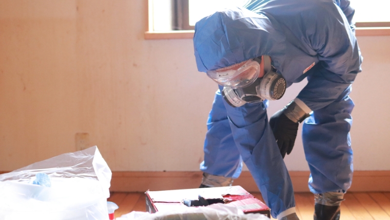 青い防護服・ゴーグル・ガスマスクを着用した特殊清掃員が床に広げたシートの上の物を確認しながら作業している様子