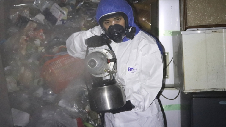 白い防護服・青いヘルメット・ガスマスクを着用した特殊清掃員がゴミが散乱した室内でステンレス製の噴霧器を使い作業している様子