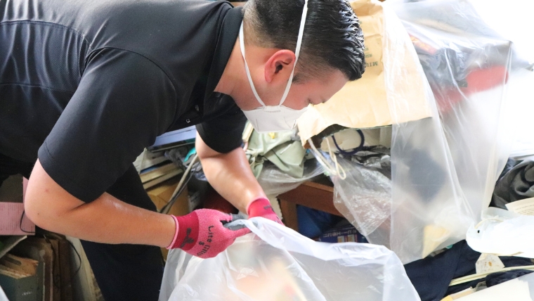 黒い服装にマスクとピンク色のグローブを着用した男性スタッフが荷物の中からビニール袋を取り出して作業している様子