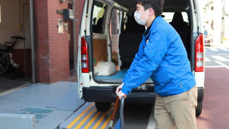 青いユニフォームとマスクを着用した男性スタッフが白いバンの荷台に荷物を積み込んでいる様子