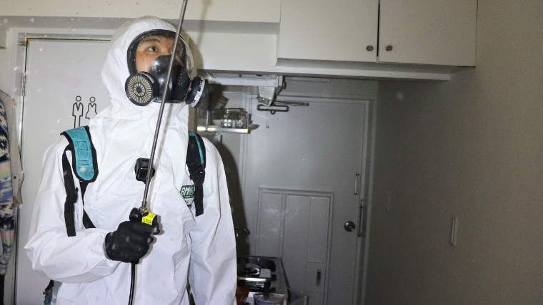 白い防護服とガスマスクを着用した特殊清掃員が背負い式の噴霧器を装着し室内でノズルを構えている様子