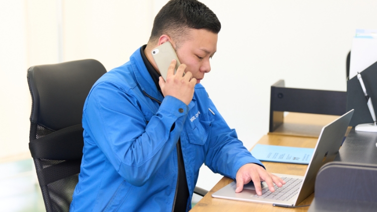 青いユニフォームを着た男性スタッフがオフィスでスマートフォンで通話しながらノートパソコンを操作している様子