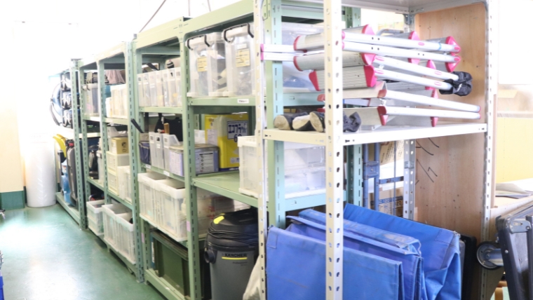 倉庫の複数の棚に清掃用具・収納ボックス・青いシートなどの資材が整然と保管されている様子