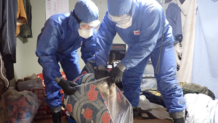 孤独死後の腐敗汚染に対応するため、防護服とマスクを着用した特殊清掃員2名が布団を透明なビニール袋に密閉梱包する様子