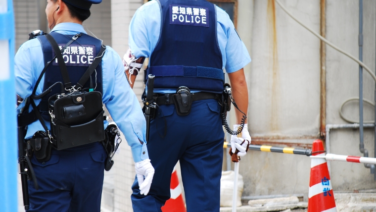 「愛知県警察」と記されたPOLICEベストを着用した警察官2名が現場へ向かう後ろ姿