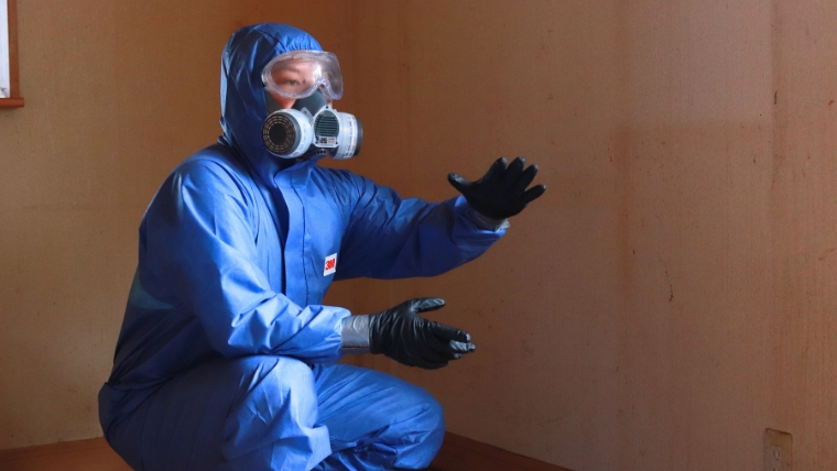 青い防護服・ゴーグル・ガスマスクを着用した特殊清掃員が室内の壁の前にしゃがみ、両手を広げて説明している様子