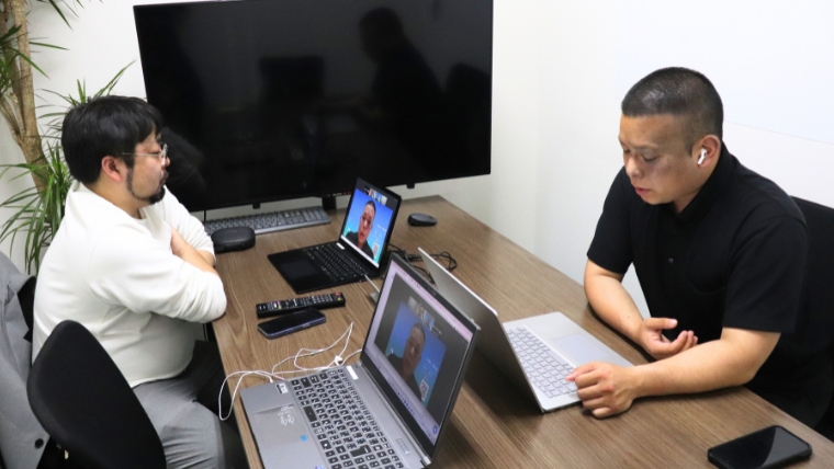 会議室のテーブルを挟んで2名の男性がそれぞれノートパソコンを前に向かい合って打ち合わせをしている様子
