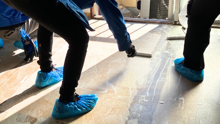 青い使い捨てシューズカバーを着用した複数のスタッフがビニールシートを敷いた床で作業している様子