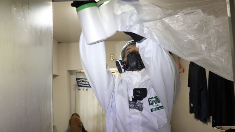 白い防護服とガスマスクを着用した特殊清掃員が室内でビニールシートを使いゾーニング作業をしている様子