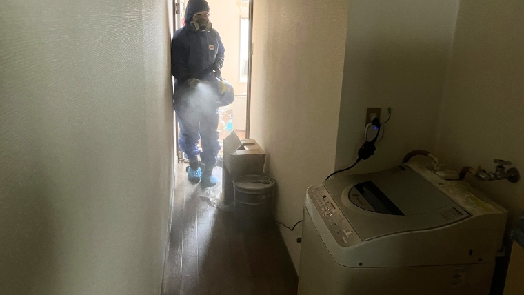 青い防護服と防毒マスクを着用した自社スタッフが、洗濯機のある室内廊下で噴霧器を使って実際に消臭・除菌作業を行っている様子