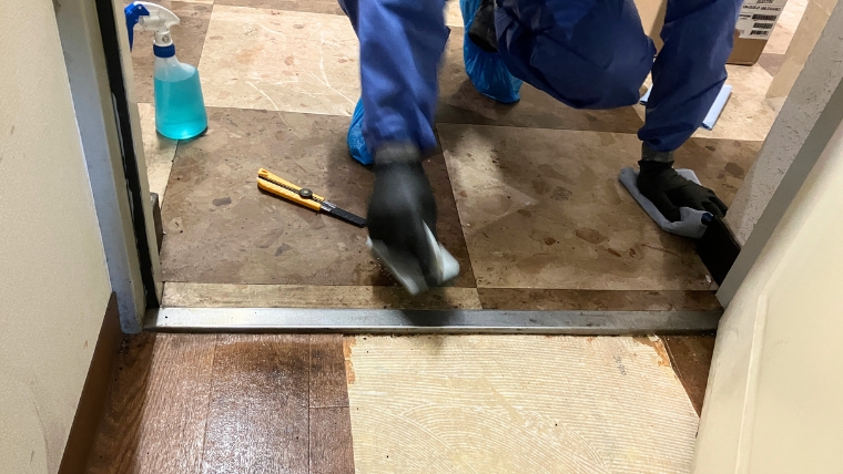 青い防護服とシューズカバーを着用した自社スタッフ2名が、汚損した玄関タイルをスポンジとカッターナイフを使って実際に清掃・除去作業を行っている様子