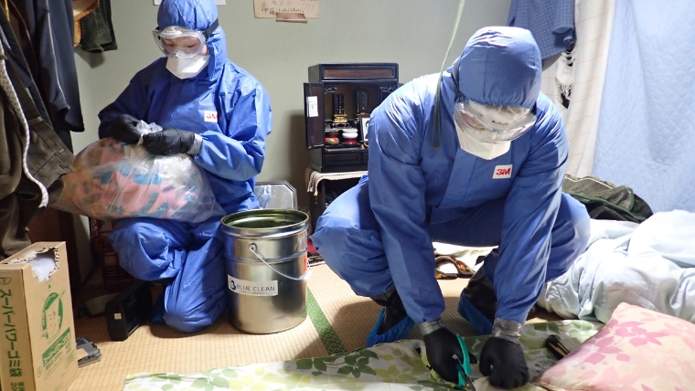 青い防護服・3M製マスク・ゴーグルを着用した自社スタッフ2名が、仏壇のある和室でブルークリーン専用資材を使って実際に特殊清掃作業を行っている様子