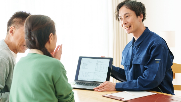 青い作業服と思われる業者担当者がノートパソコンで見積書を高齢の夫婦と思われる2名に説明しているイメージ。実家の片付けで複数業者から相見積もりを取る状況を想起させる