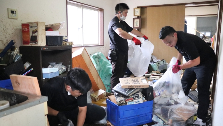 黒い作業服と赤いゴム手袋を着用したブルークリーンスタッフ3名が、荷物が散乱した実家の室内で実際に片付け作業を行っている様子
