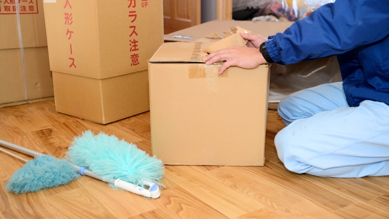 青い作業服と思われる人物が段ボール箱を床で梱包しており、傍らに清掃用ハタキが置かれているイメージ。実家の片付けを自分で進めて業者費用を抑える状況を想起させる