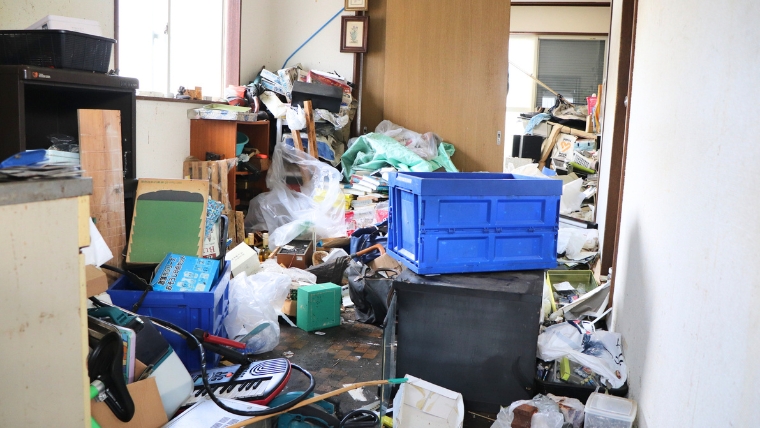 自社スタッフが撮影した実家の片付け現場。家具・書籍・雑貨・ゴミが室内全体に堆積し、業者依頼が必要な状態の実際の現場写真