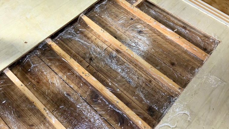 自社スタッフが撮影した床材剥離後の根太・床下地。ペットの尿が長期間浸透し黒く変色した実際の現場写真。薬剤塗布後にラップで養生している状態