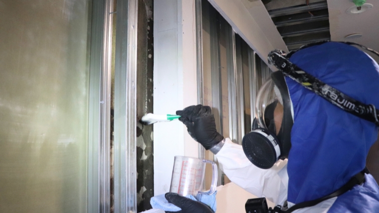 青い防護服と防毒マスクを着用した自社スタッフが、計量カップに入れた薬剤をハケで壁面のカビ汚染部に実際に塗布している様子