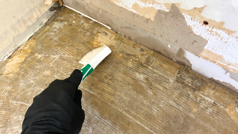 黒いゴム手袋を着用した自社スタッフが、床材が剥離した下地にスクレーパーで薬剤を塗布し臭いの根本を実際に除去している様子
