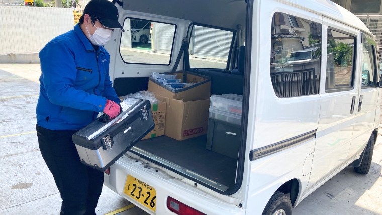 青い作業服とピンクのゴム手袋を着用したブルークリーンスタッフが、車両のトランクから特殊清掃用工具・資材を取り出し現場へ向かう準備をしている様子