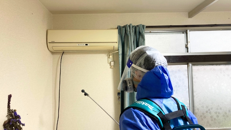 青い防護服・フェイスシールド・防毒マスクを着用した自社スタッフが、makita製背負い式散布機を装着しエアコン周辺に消臭・除菌剤を実際に散布している様子