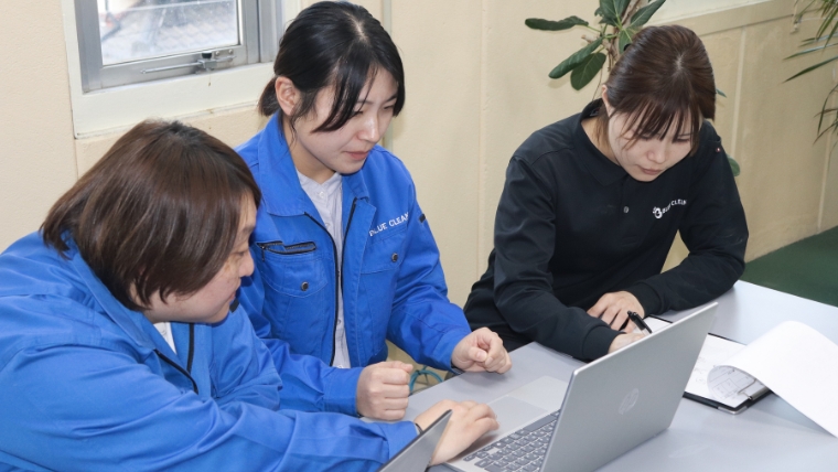 BLUE CLEANロゴ入り作業服を着用したブルークリーン女性スタッフ3名がノートパソコンを囲んで打ち合わせをしている様子