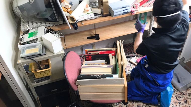 紫のゴム手袋を着用した自社スタッフが、書籍・家電・雑貨が散乱した室内で実際に片付け作業を行っている様子