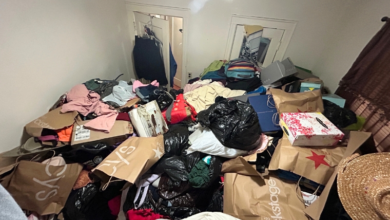 自社スタッフが撮影した衣類・ショッピングバッグ・ゴミ袋が床一面に山積みになった室内。片付け業者への依頼が必要な状態の実際の現場写真